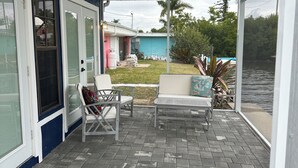 Un patio couvert avec un ventilateur de plafond, des sièges d’extérieur et une vue sur un bâtiment bleu et sur l’eau.
