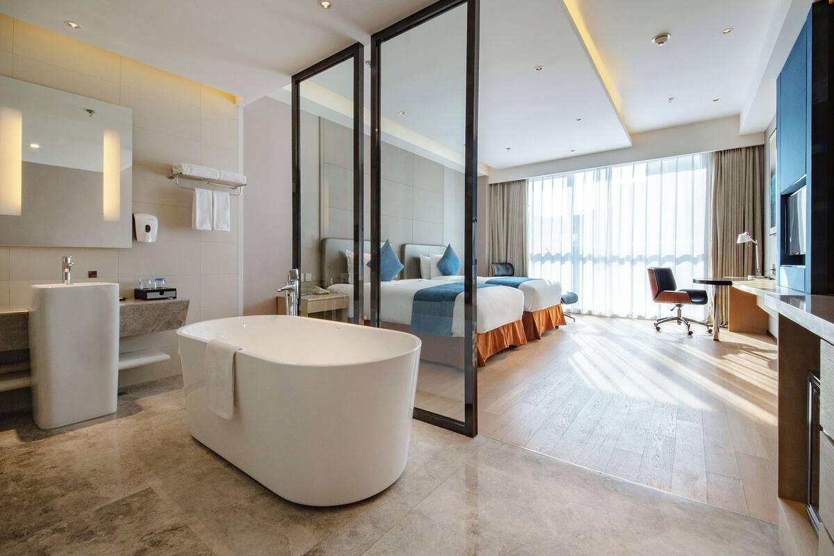Una camera d'albergo moderna con una vasca da bagno spaziosa, una zona doccia separata, un letto con vista e una scrivania con sedia.