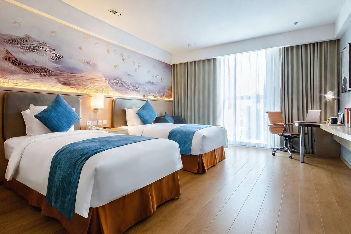 Una camera d'albergo con due letti, una scrivania, una sedia e un'ampia finestra con tende.
