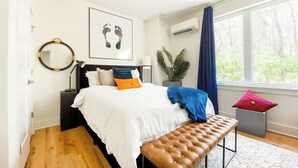 Ein Schlafzimmer mit Bett, rundem Spiegel, einem Wandbild mit Fußabdruck, einer Topfpflanze und einem Fenster mit Vorhängen.