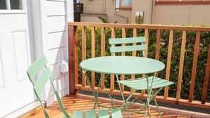 Une terrasse en bois avec une table et des chaises en métal vert, une porte blanche et un bâtiment beige en arrière-plan.