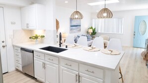 A modern kitchen with white cabinetry, a central island with a sink, pendant lights, and a dining area with chairs and a table. - The Orkney - A Modern Coastal Cardiff Residence (Encinitas)
