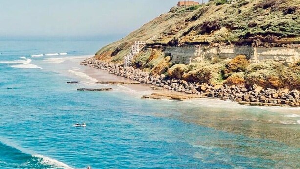 A coastal landscape with a rocky beach, clear blue water, and a person surfing. - The Orkney - A Modern Coastal Cardiff Residence (Encinitas)