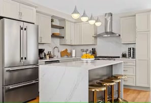 A modern kitchen with white cabinets, stainless steel appliances, a marble island, and bar stools. - SkyDeck Stylish Stay (Somerville)