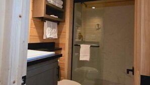 Une salle de bain rustique, aménagée dans une cabane en bois, avec des toilettes, une douche en verre et des étagères en bois où sont rangées les serviettes.