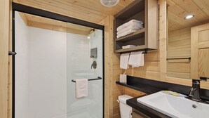 A modern bathroom with a glass shower enclosure, a white toilet, a sink with a black countertop, and wooden shelves with towels. - Cozy Tiny Home with Loft Sleeps 6 (Page)