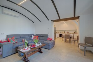 Ein modernes Wohnzimmer mit einem grauen Sofa, einem Holztisch und einem Essbereich, im Hintergrund ist eine Küche zu sehen.