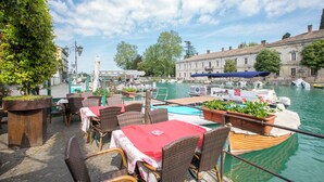 An outdoor dining area with wicker furniture and a red tablecloth, overlooking a canal with boats and a building in the background. - Peschiera few steps from the lake Garden, Terrace, & AC, Peschiera del Garda, Italy (Peschiera del Garda)