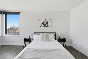 Een slaapkamer met een groot bed, twee nachtkastjes, een raam met uitzicht op de stad, en een schilderij aan de muur.