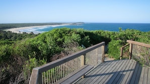 A wooden deck with a railing overlooking a coastal landscape with a beach and ocean.