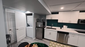 A compact kitchen with white cabinets, a stainless steel refrigerator, a microwave, a dishwasher, and a small dining area with a chair and table. - Modern Private Suite with 100 inch TV Quiet Oshawa (Oshawa)