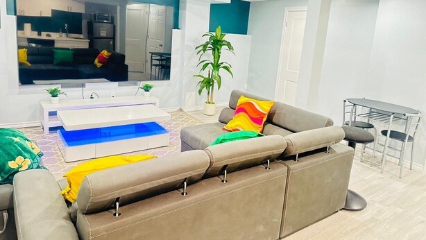 A modern living room with a sofa, a coffee table, a TV, and a kitchen area in the background. - Modern Private Suite with 100 inch TV Quiet Oshawa (Oshawa)