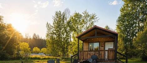 A wooden cabin with a porch, surrounded by a grassy field and trees.