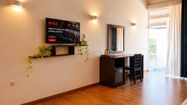 Habitación de hotel con televisión de pantalla plana montada en la pared, un escritorio con una silla, un mueble pequeño y un ventanal con cortinas.