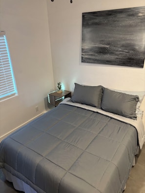Ein Schlafzimmer mit einem großen Bett, einem Nachttisch mit Lampe, einem Wandbild und einem Fenster mit Jalousien.