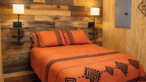 A bed with an orange bedspread, two wall-mounted lamps, a patterned rug, and a wooden wall panel. - Sweet county cabin in the cozy county (Jennings)