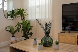 Un salon avec une table en bois, des plantes en pot, un vase vert, un téléviseur et une fenêtre ornée de rideaux légers.