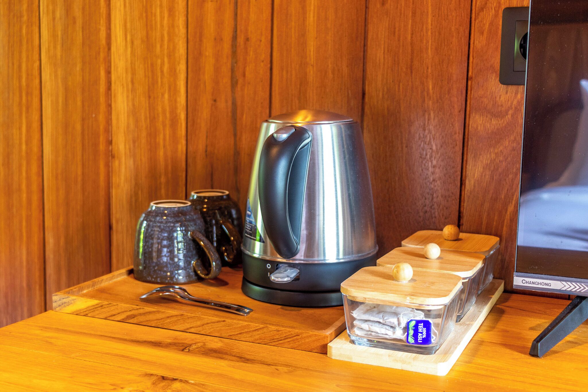 A kitchen area with a stainless steel kettle, a ceramic mug, a wooden tray with containers, and a digital clock.