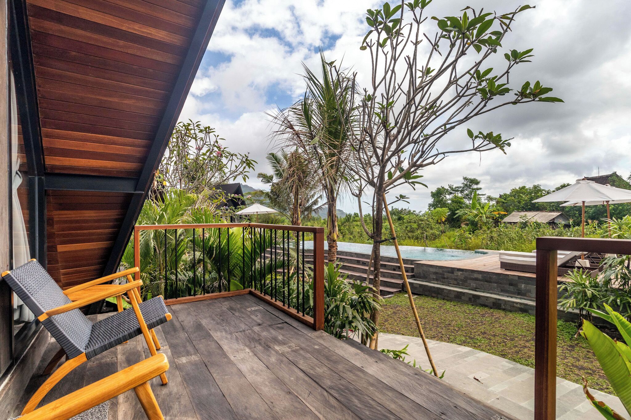 Terraza de madera con una tumbona, piscina y abundante vegetación.