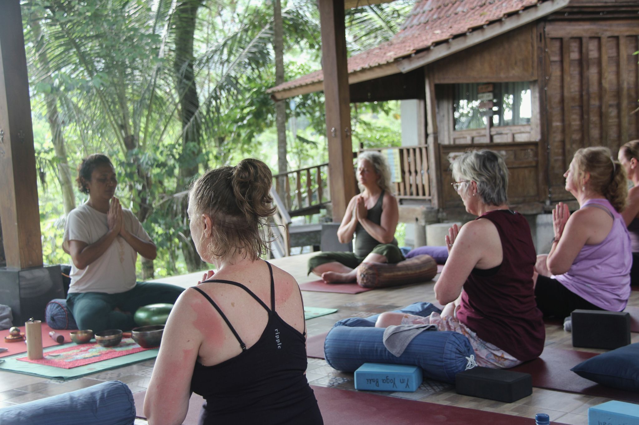 A group of people sitting on mats in a covered outdoor area with wooden structures and greenery in the background.