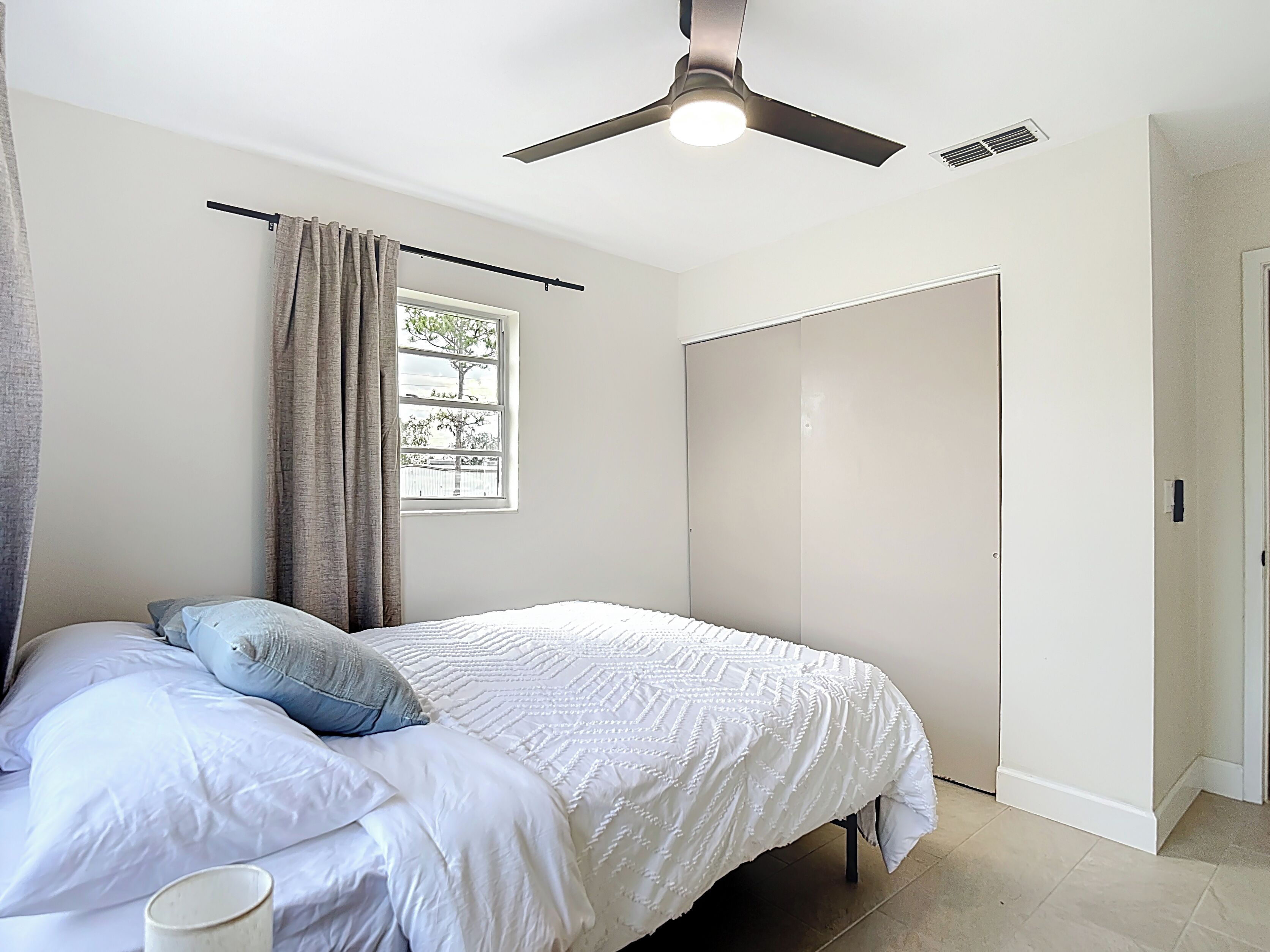 A bedroom with a bed, a ceiling fan, a window with curtains, and a door.