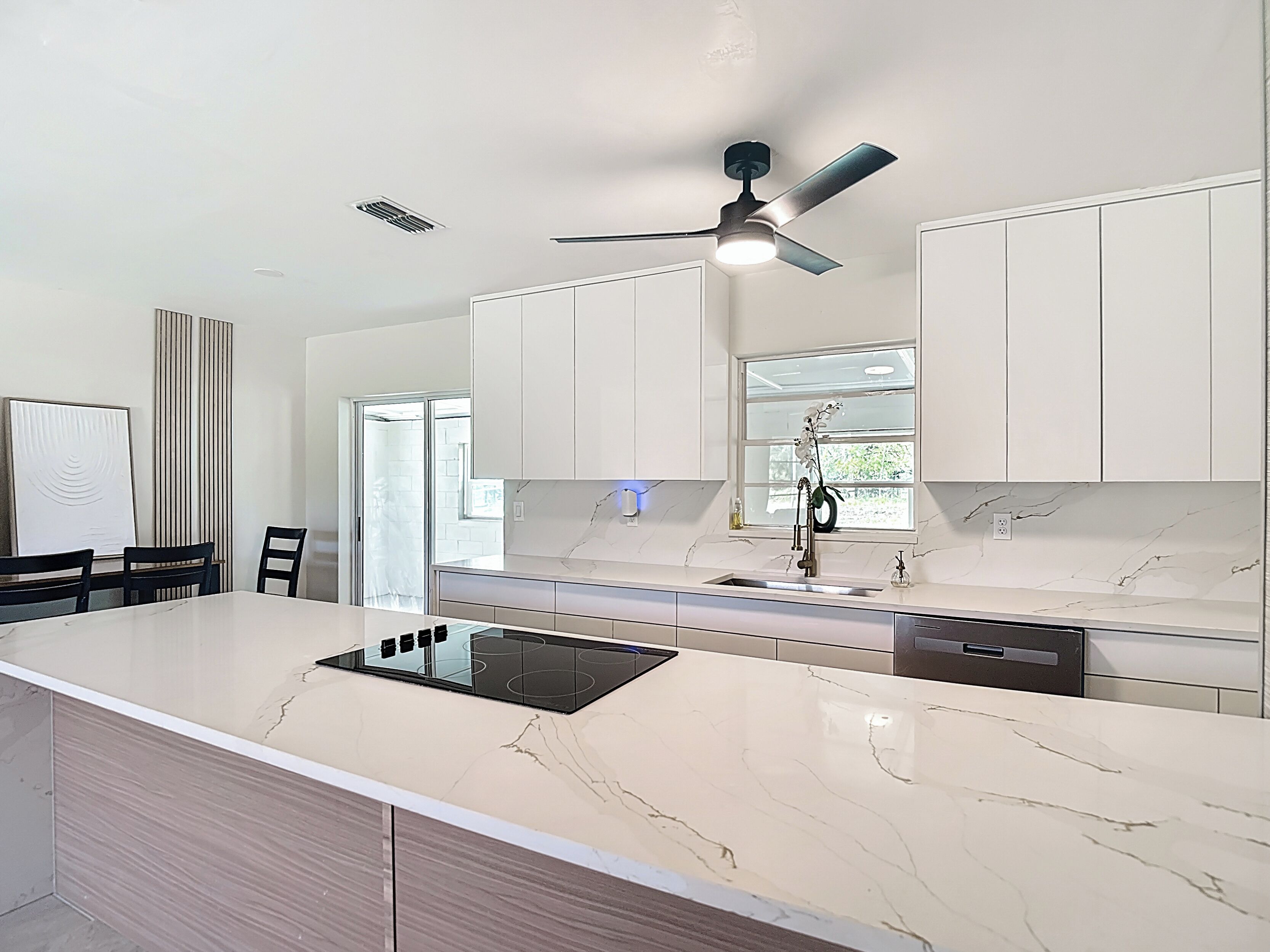 Een moderne keuken met witte kasten, een centraal eiland met een inductiekookplaat, een plafondventilator en een raam met uitzicht.