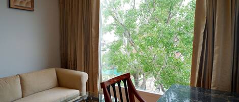 A room with a sofa, a dining table, and a chair. There is a window with curtains, and a tree is visible outside.