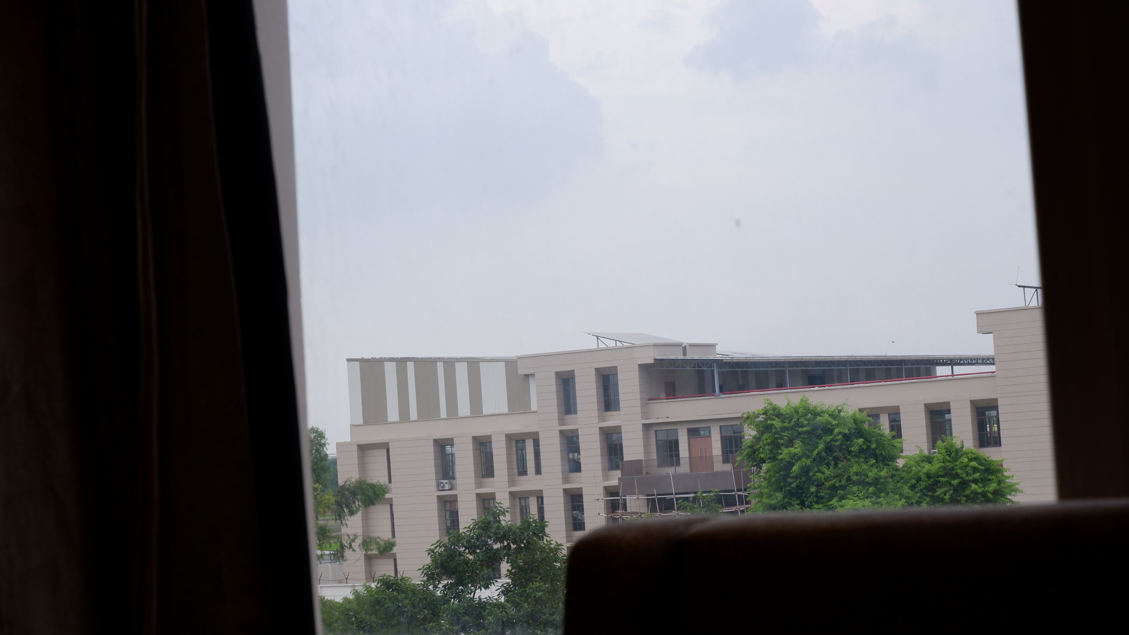 A view of a building with multiple floors and a flat roof, seen through a window with curtains.