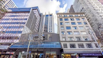 A modern urban street with tall buildings, including a glass-fronted structure and a building with a classical facade.