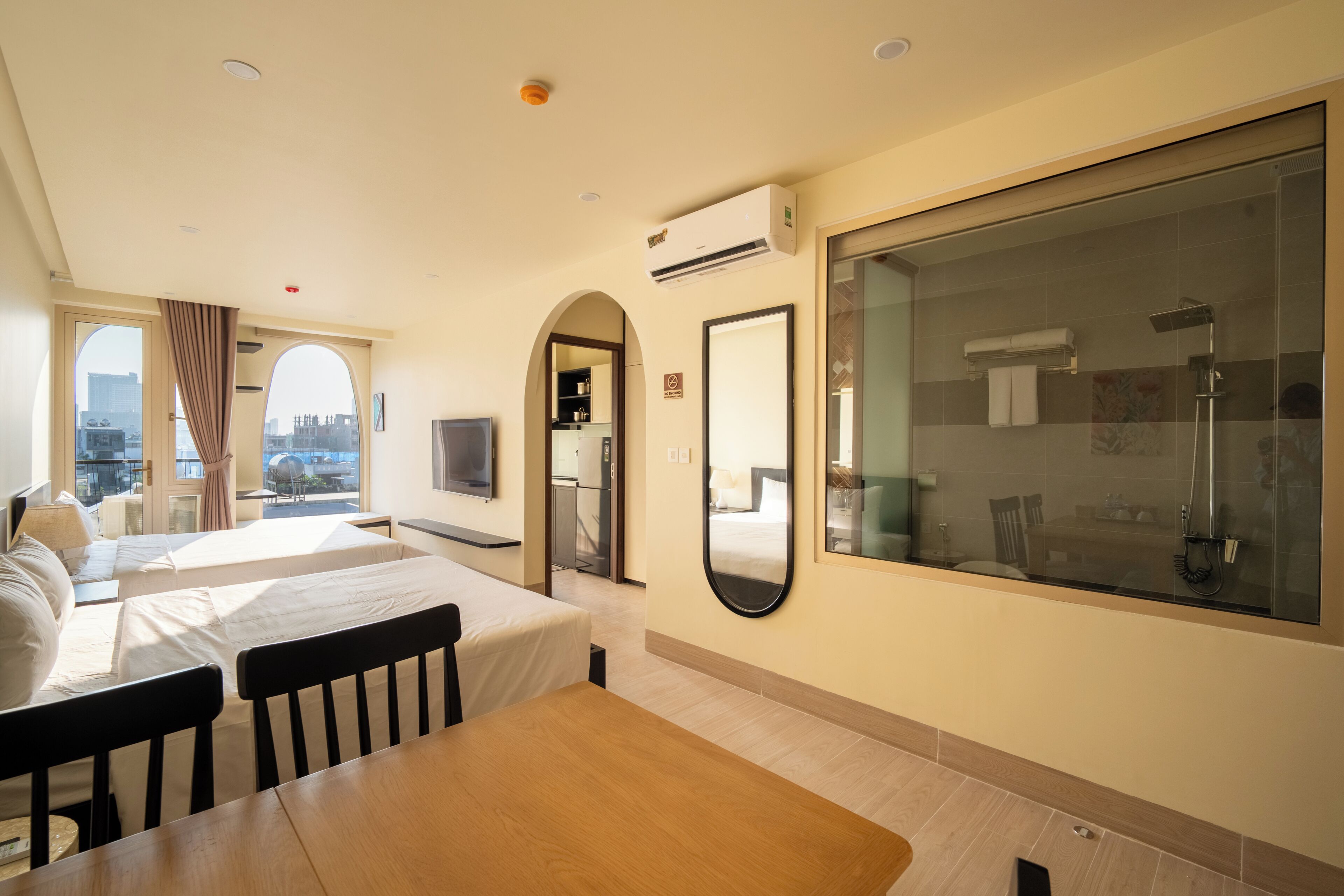 A hotel room with two beds, a dining table, a TV, a large window, and a shower area.