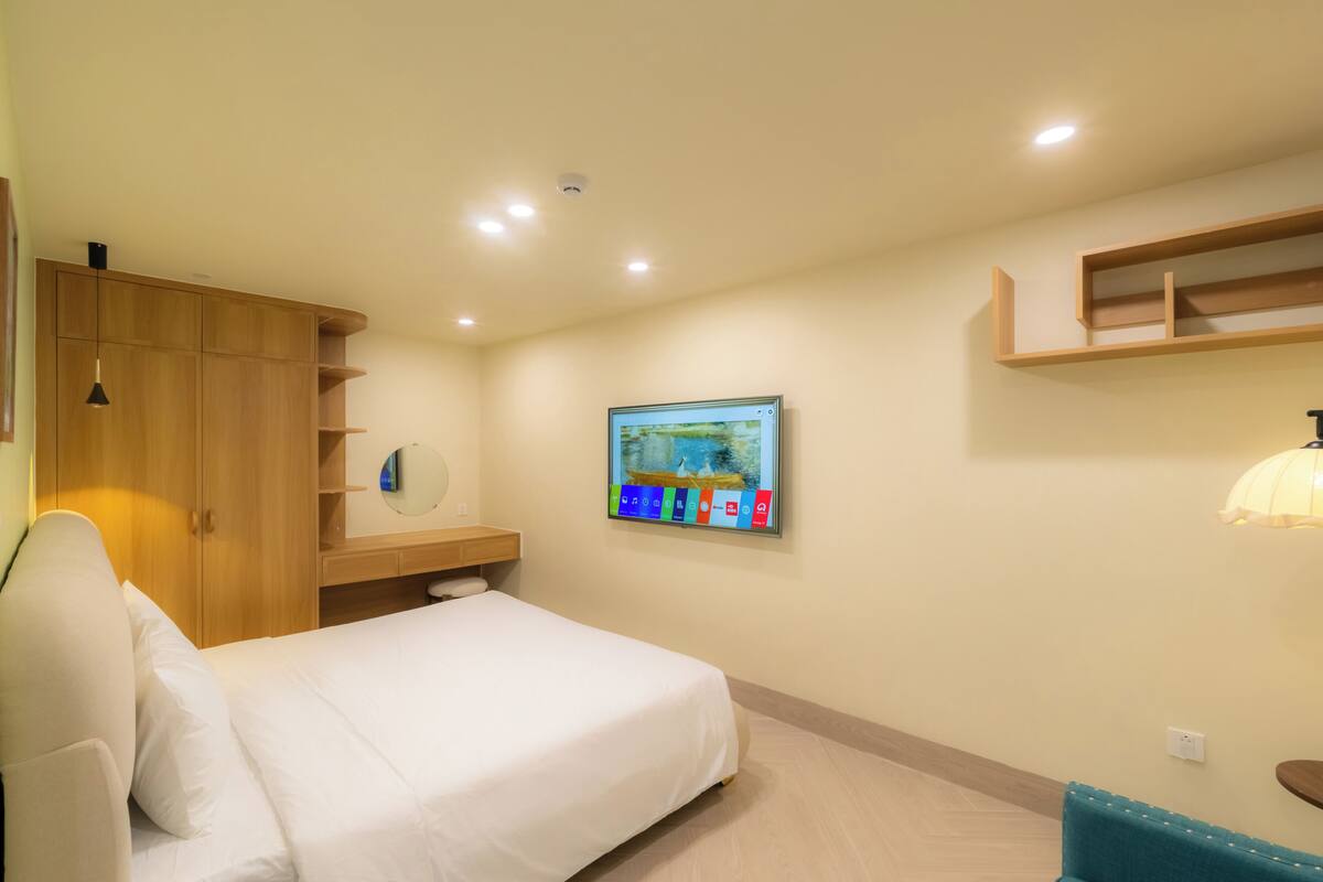 A bedroom with a bed, wooden wardrobe, wall-mounted TV, and a shelf.