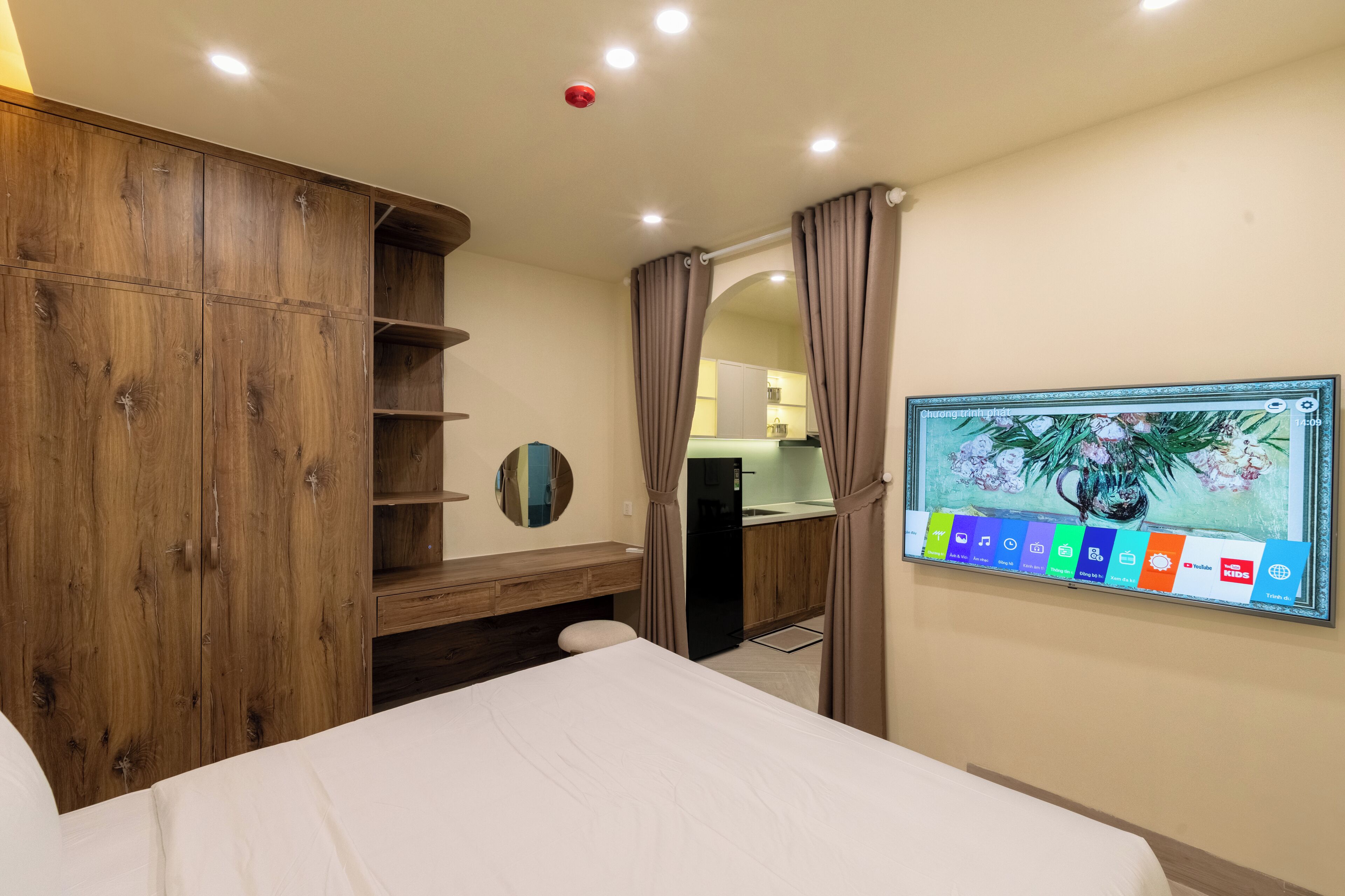 A hotel room with a bed, wooden wardrobe, a mirror, a TV mounted on the wall, and a kitchenette area.