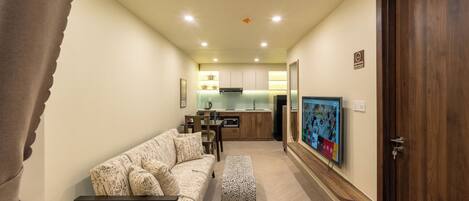 Una sala de estar con un sofá, una mesa de centro, un televisor montado en la pared y una cocina al fondo.