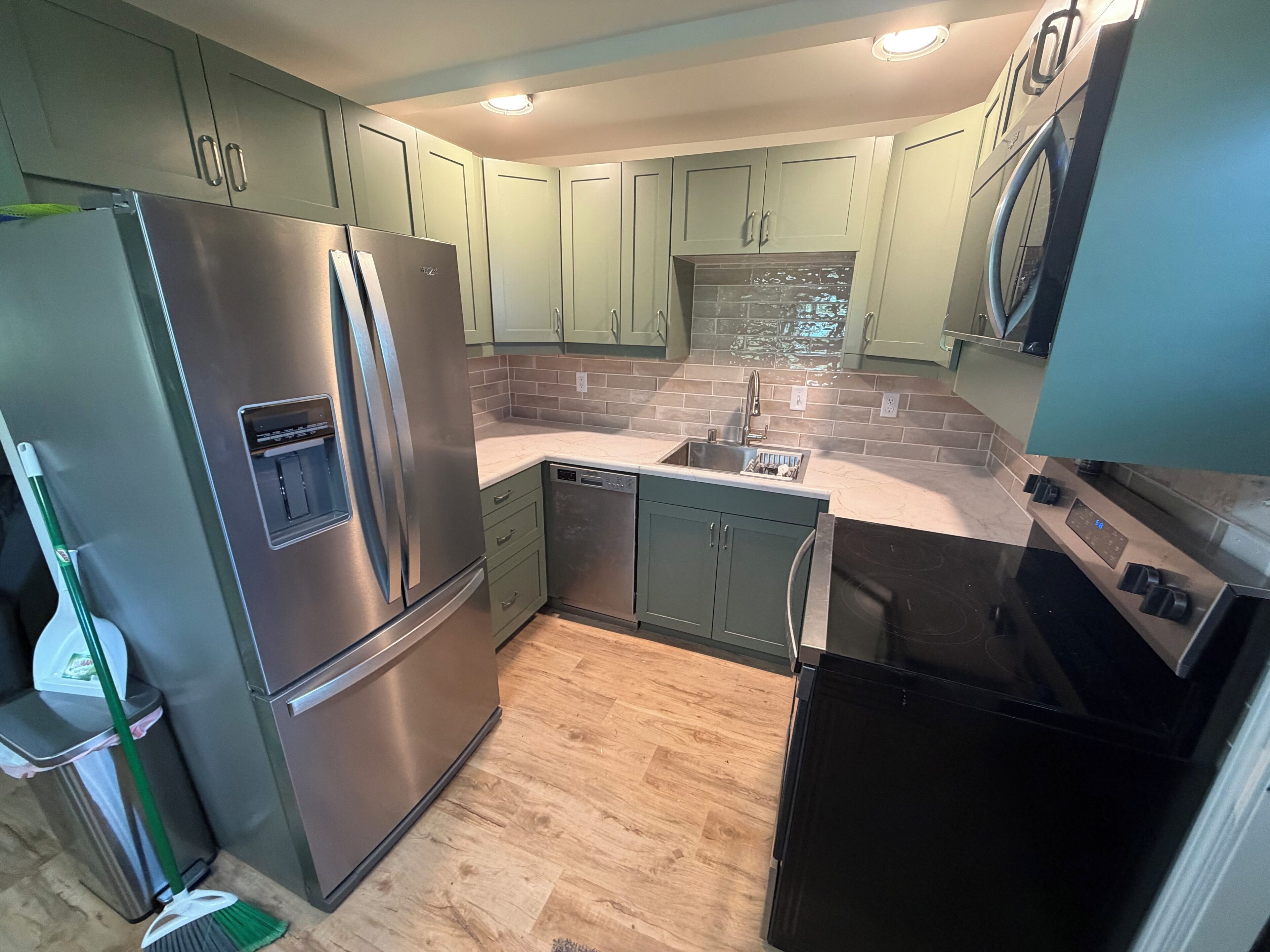 A modern kitchen with stainless steel appliances, green cabinets, a tiled backsplash, and a wooden floor.