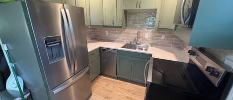 A modern kitchen with stainless steel appliances, green cabinets, a tiled backsplash, and a wooden floor.