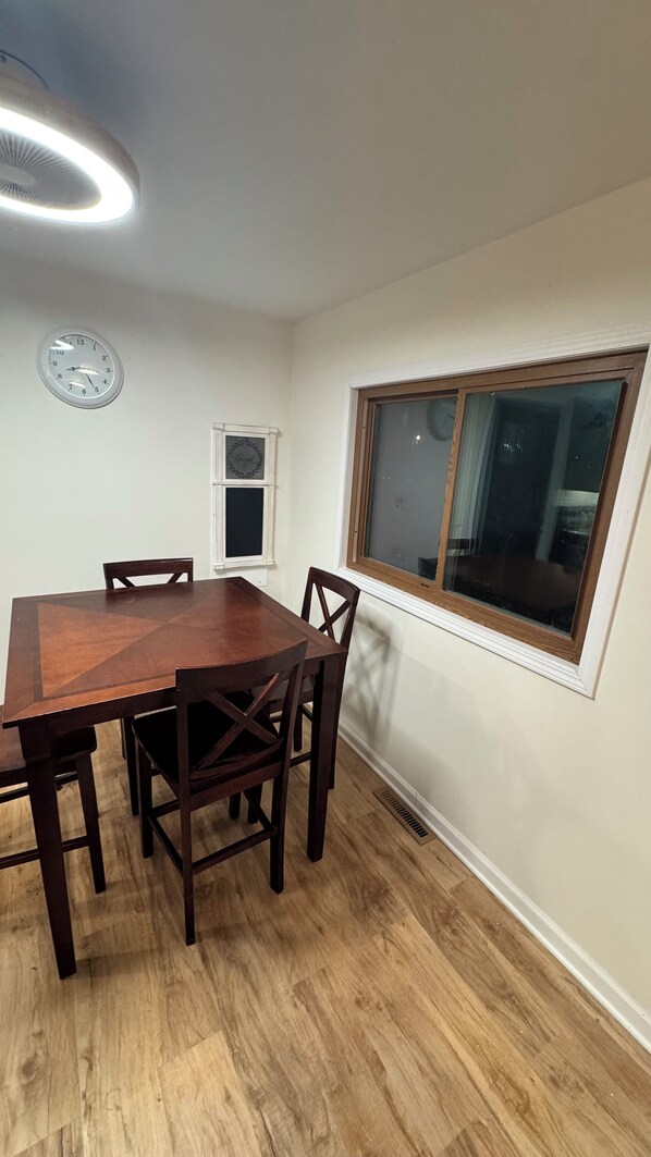 Ein Essbereich mit einem Holztisch, Stühlen, einer Wanduhr, einem Fenster und einer Deckenleuchte.