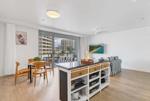 A modern living room with a dining area, a kitchen island, and a large window with a city view. - Belle Escapes 403 Harbour Lights (Cairns)
