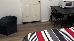 A room with a white door, a small table with a microwave, a black ottoman, and a striped bedspread. - Serene casita with AC in beautiful Coachella perfect for getaway (Coachella)