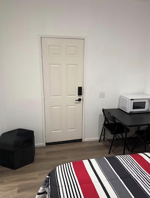 A room with a white door, a small table with a microwave, a black ottoman, and a striped bedspread. - Serene casita with AC in beautiful Coachella perfect for getaway (Coachella)