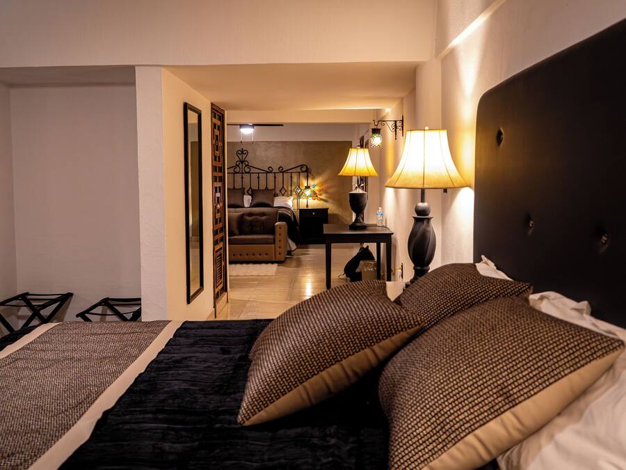 Ett hotellrum med en säng, sängbord, en lampa och utsikt in i det intilliggande rummet.