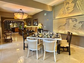 Un comedor con una mesa grande dispuesta para una comida, rodeada de sillas. Hay un candelabro sobre la mesa y una ventana con persianas a la derecha.