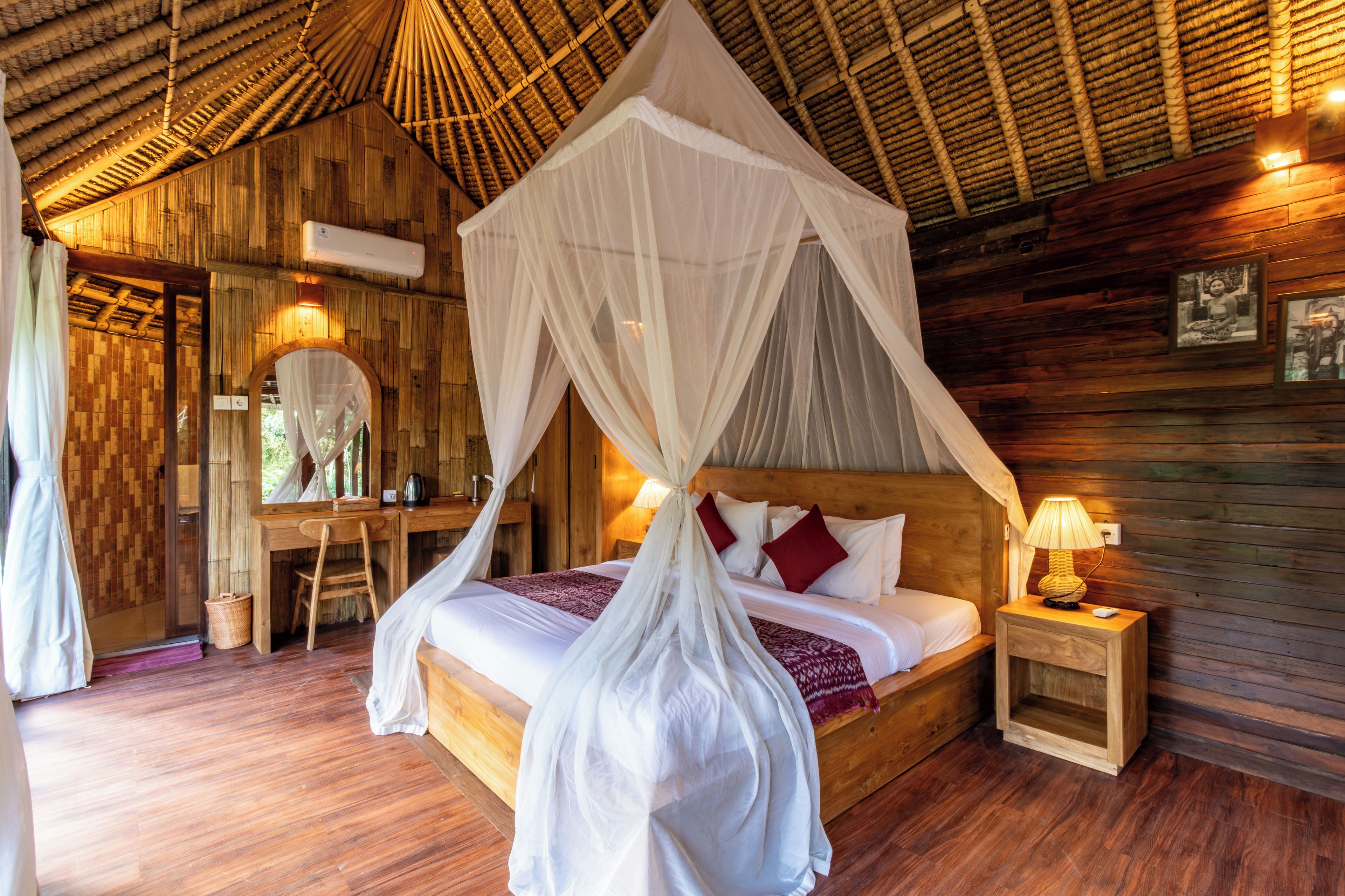Een kamer met een houten plafond, een bed met wit beddengoed en een hemelbed, een bureau met stoel, en een nachtkastje met lamp.
