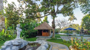 A garden with a statue, a thatched-roof structure, a pond, and a statue by the pool.