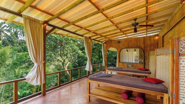 A wooden room with two massage tables, a ceiling fan, and a view of lush greenery.