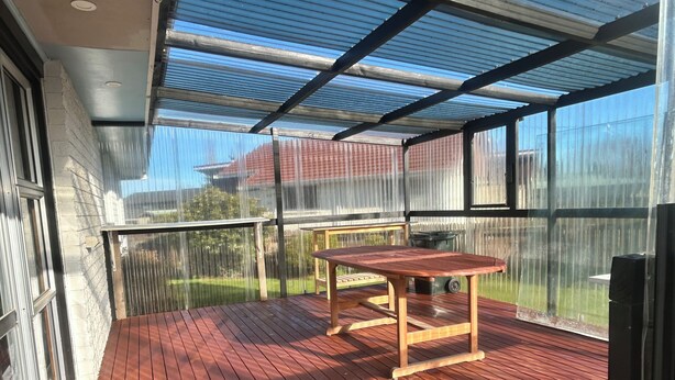 Un espacio al aire libre cubierto con una mesa y sillas de madera, una barandilla metálica y vistas a una casa a través de una malla.