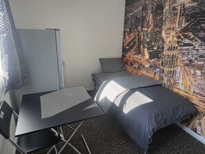 Una habitación pequeña con cama, un escritorio plegable y un cuadro con vista urbana.