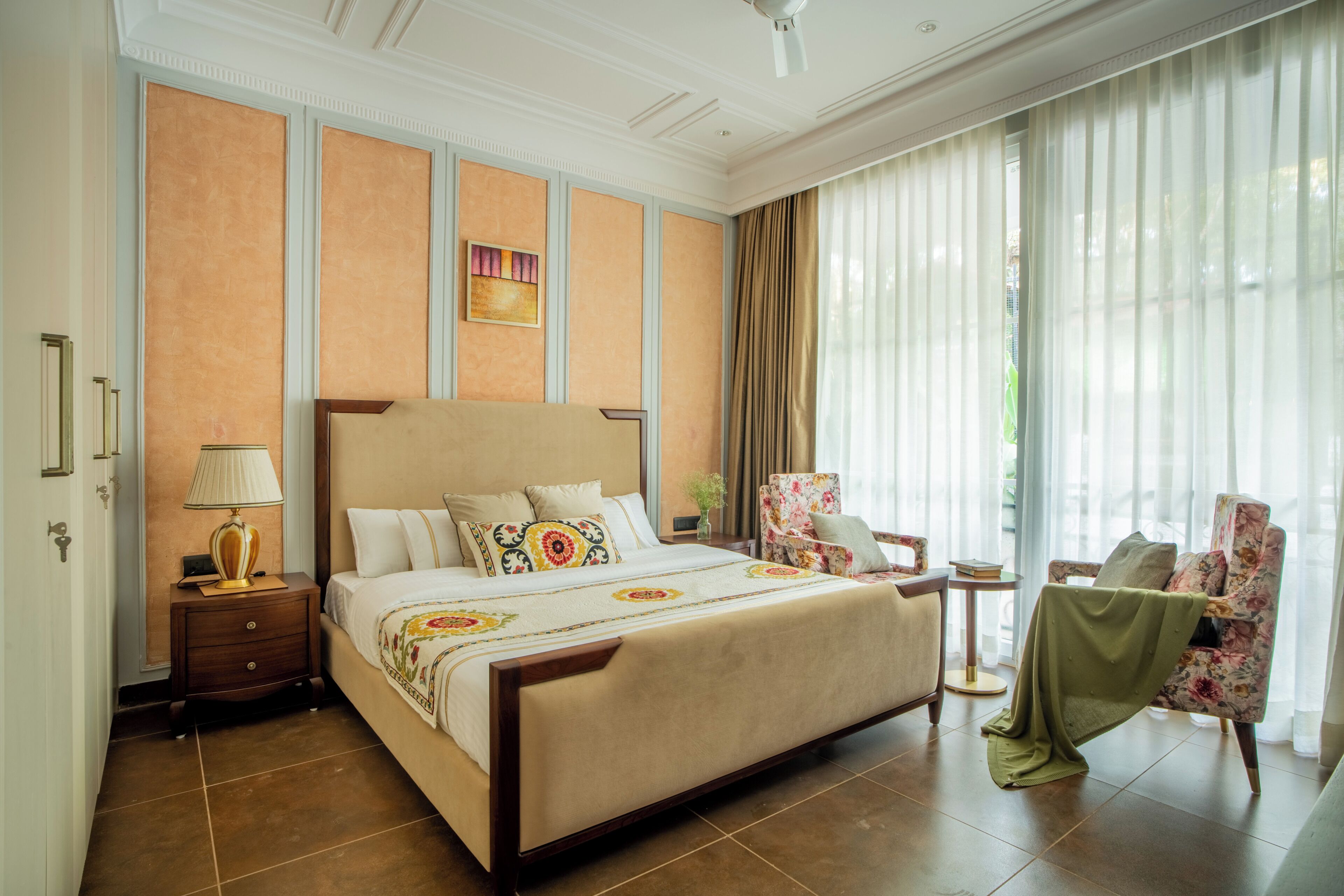 A hotel room with a large bed, a nightstand, a chair, and a view of the outdoors through sheer curtains.