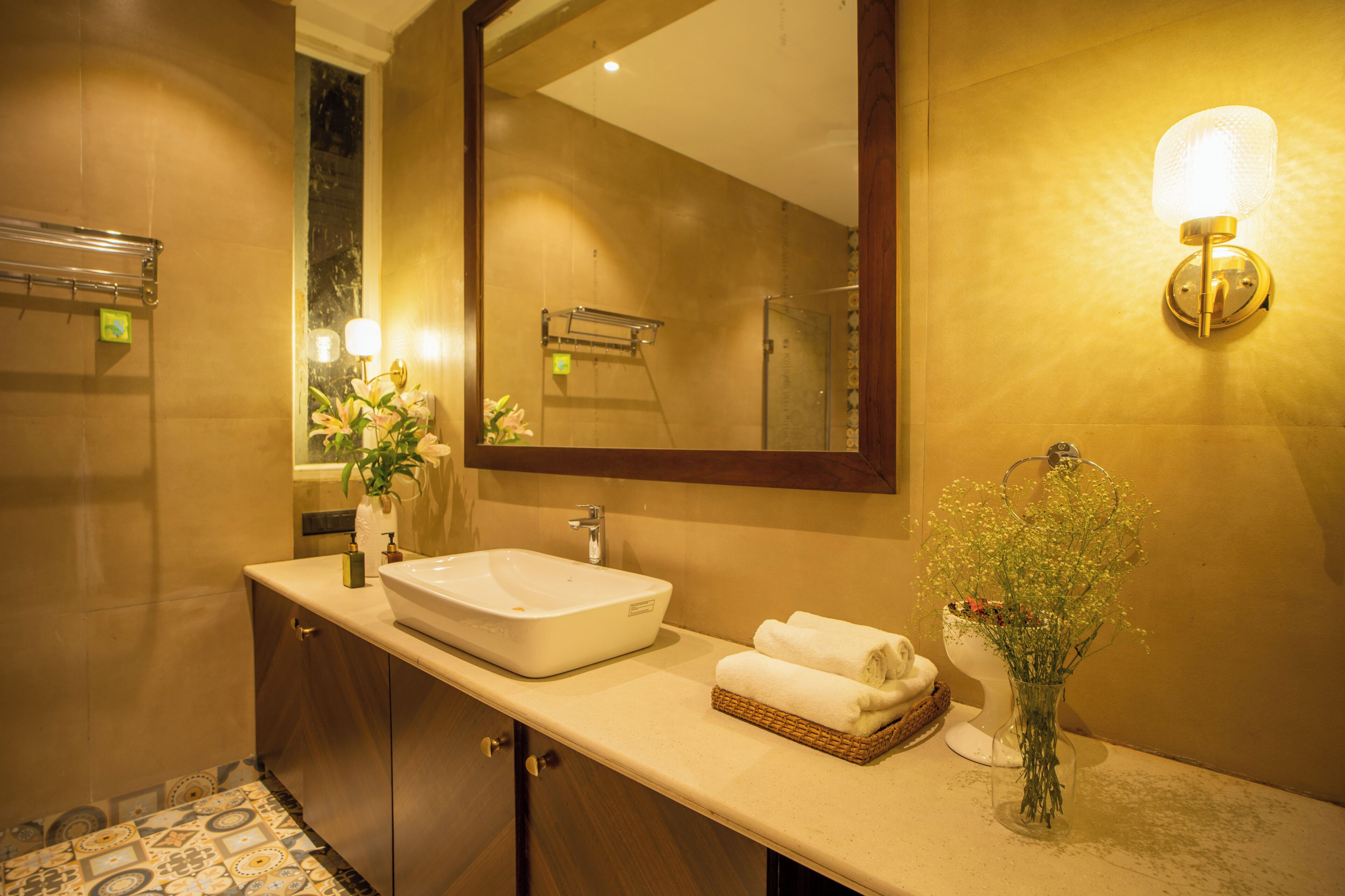 Un baño con un espejo grande, un lavamanos, un florero con flores y un perchero para toallas.