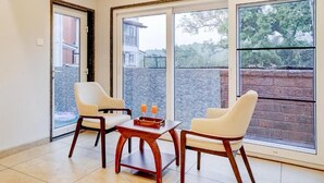 Una habitación con dos sillas, una mesita pequeña y amplias ventanas que dan a un muro de ladrillo y árboles.