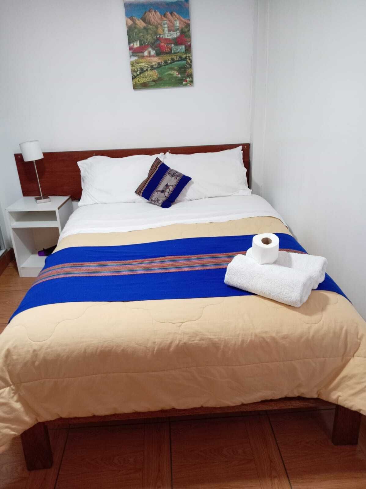 A neatly made bed with a blue and beige bedspread, a white pillow, and a rolled white towel.